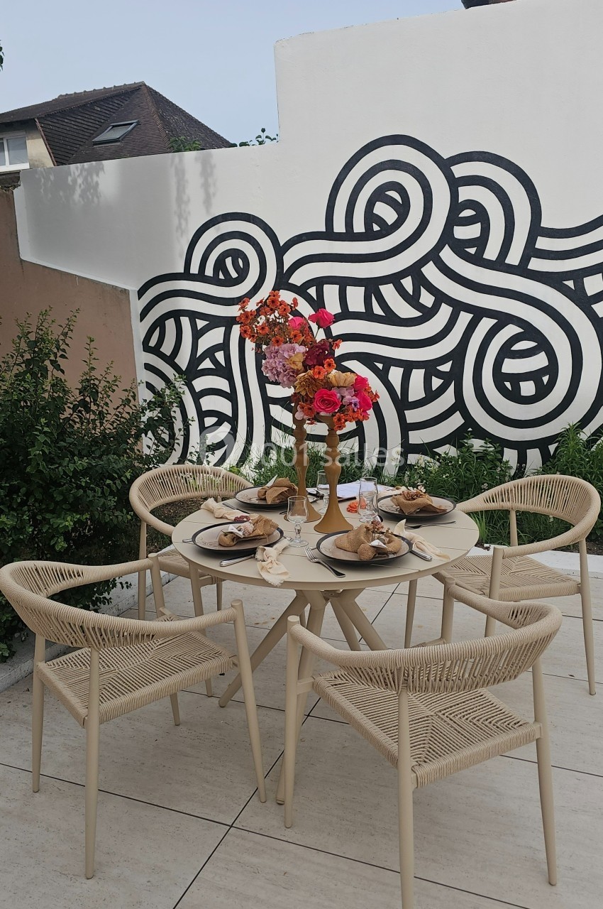 Table de jardin beige avec chaises assorties, décorée de fleurs colorées et dressée pour un repas, devant une fresque murale.