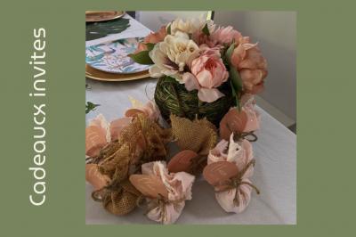 Arrangement floral avec des fleurs roses et blanches, entouré de petits sachets cadeaux en tissu et jute sur une table.
