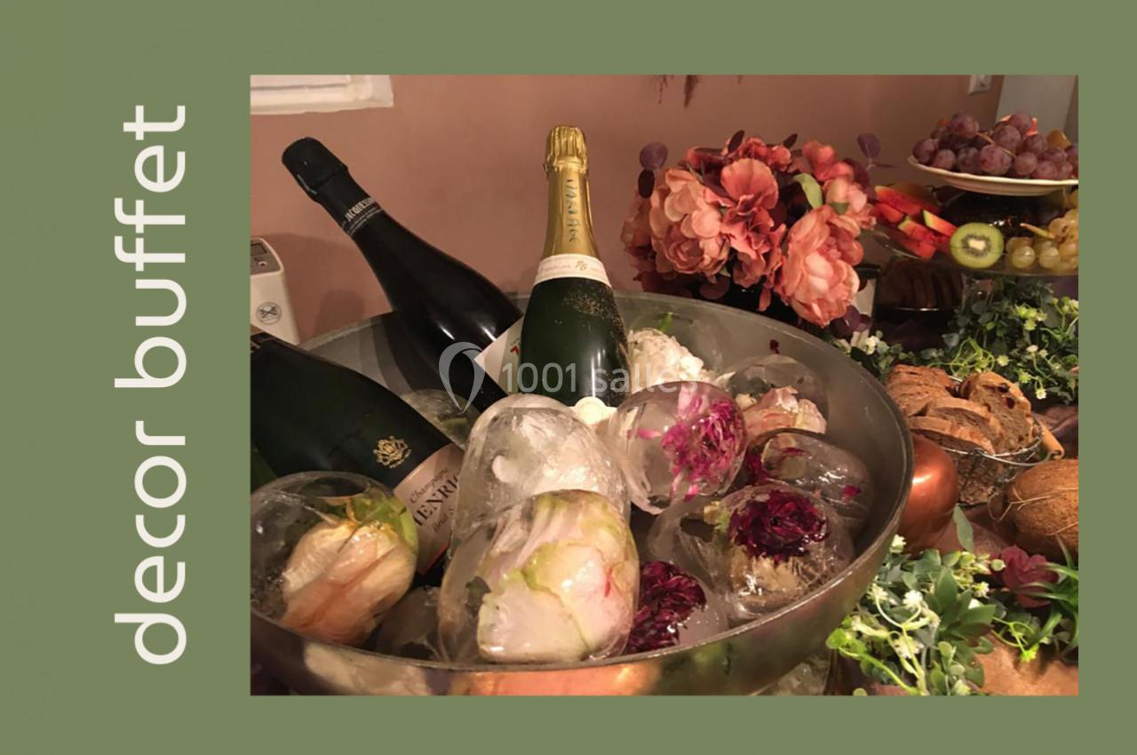 Bouteilles de champagne dans un seau rempli de glace avec des fleurs, entourées de fruits et décorations végétales.