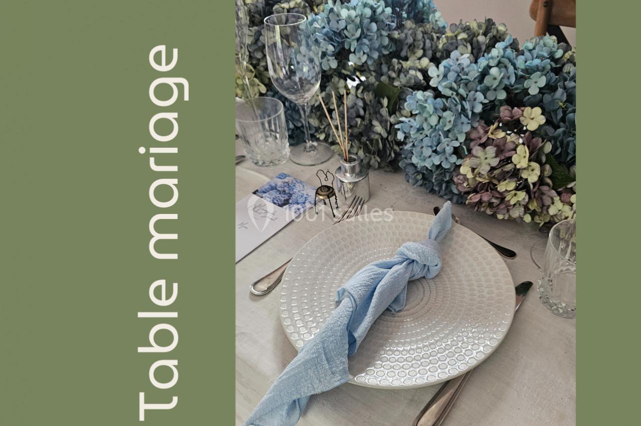 Table élégamment dressée avec assiette blanche, serviette bleue nouée et décoration florale en hortensias.