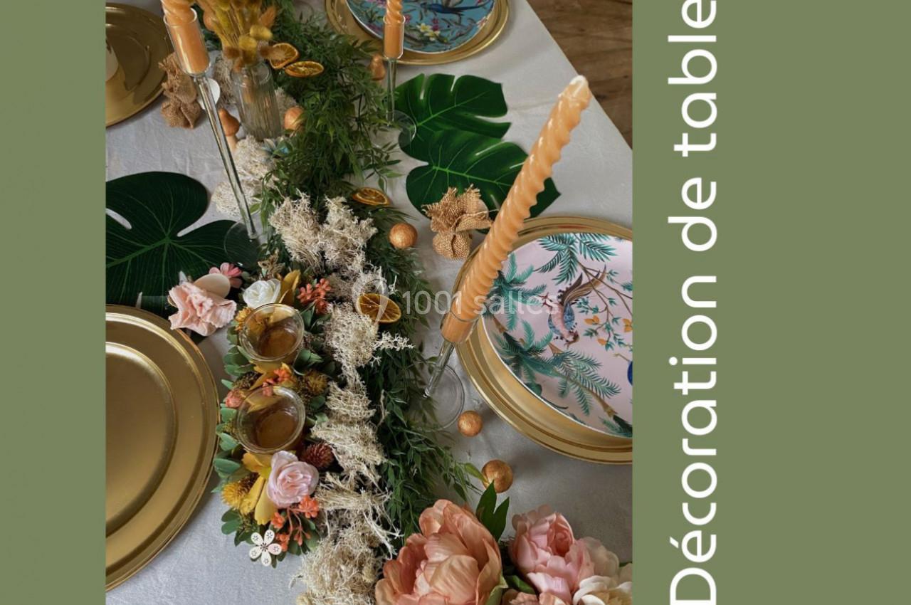 Centre de table végétal avec fleurs, bougies torsadées, assiettes dorées et motifs tropicaux sur une nappe blanche.