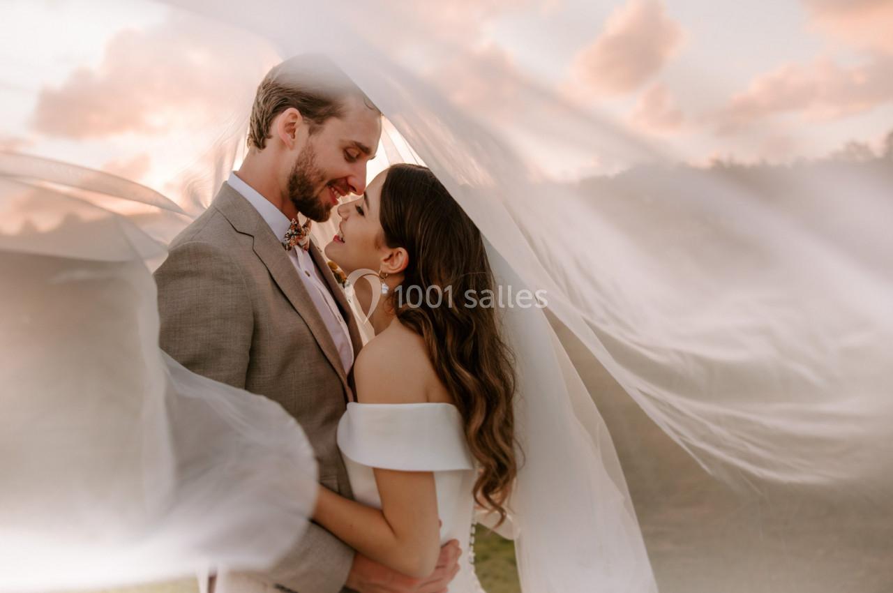 Un couple en tenue de mariage s'enlace sous un voile flottant, avec un ciel nuageux en arrière-plan.
