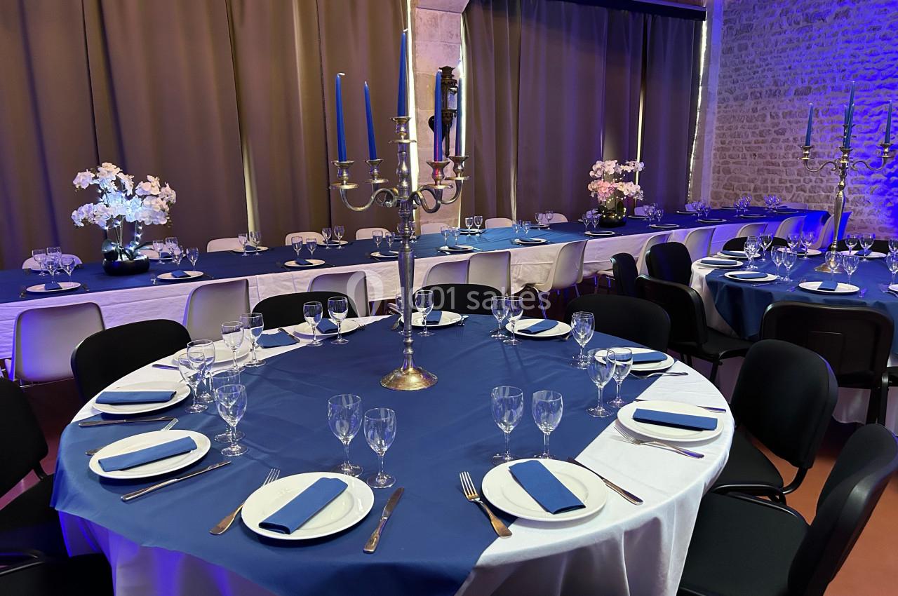 Salle décorée pour un événement avec tables rondes dressées, nappes bleues, chandeliers et éclairage tamisé.
