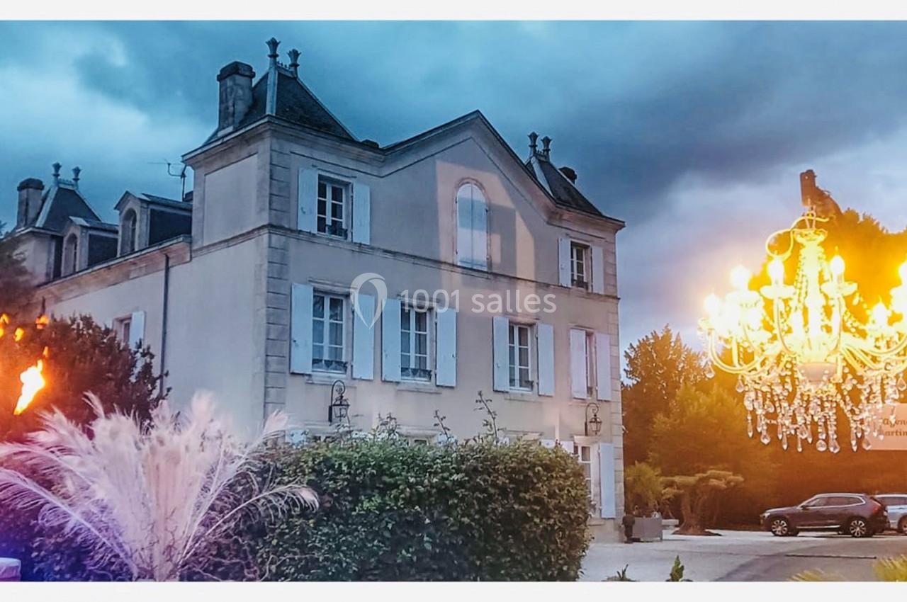 Manoir ancien entouré de végétation, éclairé par un lustre extérieur et des torches, sous un ciel nuageux.