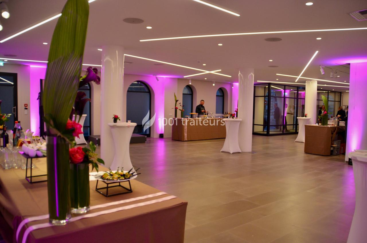Salle de réception moderne avec éclairage rose, tables hautes et buffet décoré de fleurs.