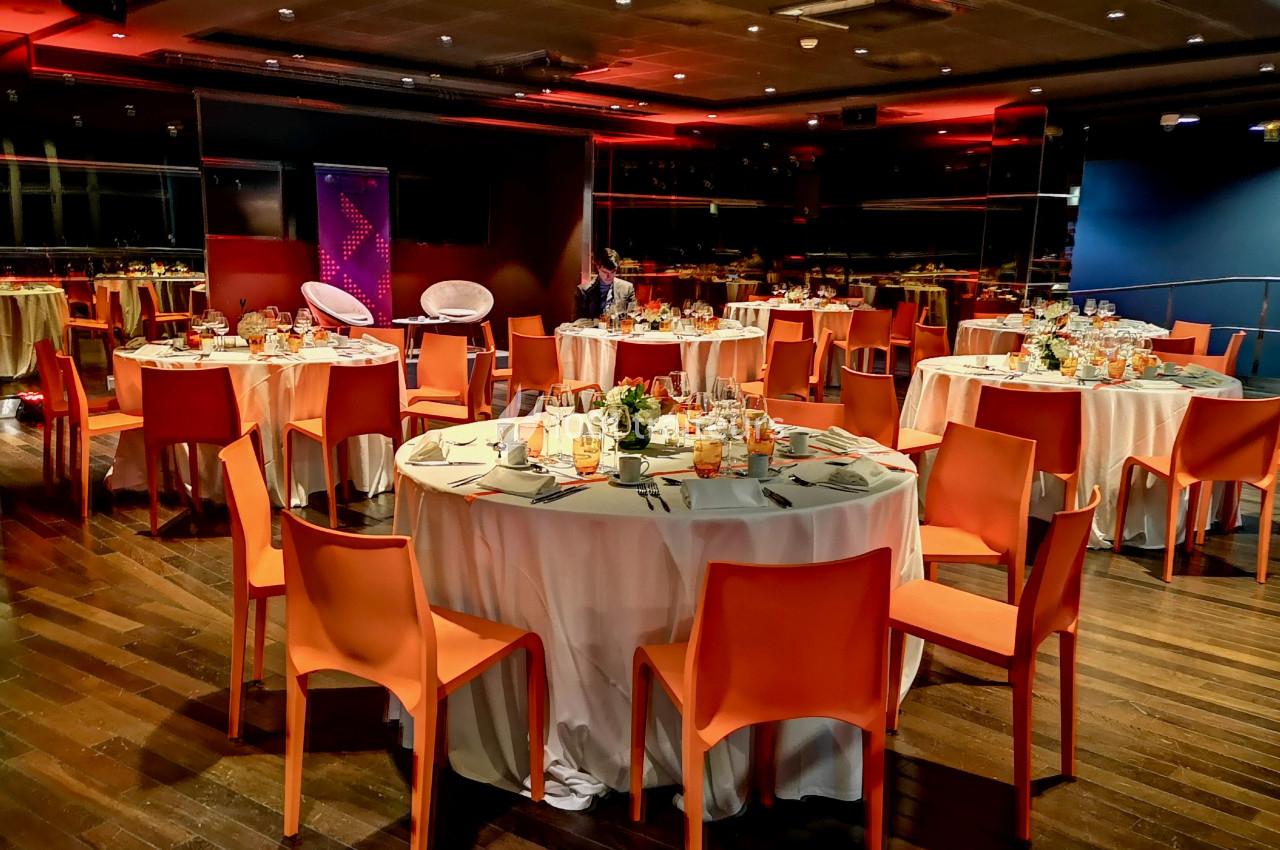Salle de réception avec des tables rondes dressées, nappes blanches, chaises orange et éclairage tamisé.