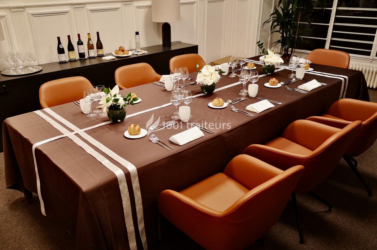 Table élégamment dressée avec nappe marron, vaisselle, verres, fleurs blanches et chaises orange dans une salle lumineuse.