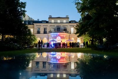 Miniature Traiteur Saint-Cloud (Hauts-de-Seine) - Aux Jardins des Sens #51 Façade illuminée d'un bâtiment élégant au crépuscule, avec un jardin et des invités rassemblés devant.