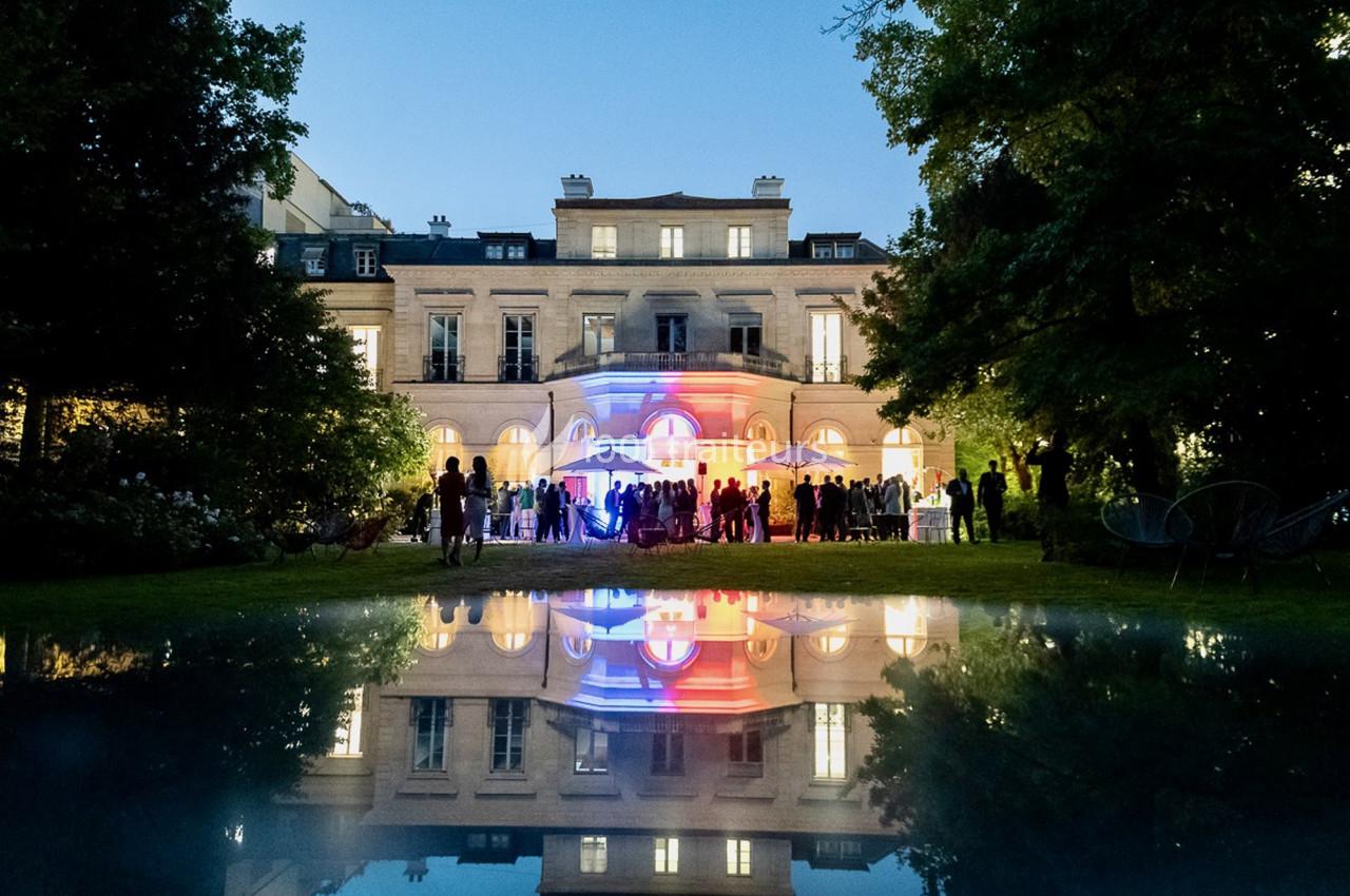 Façade illuminée d'un bâtiment élégant au crépuscule, avec un jardin et des invités rassemblés devant.