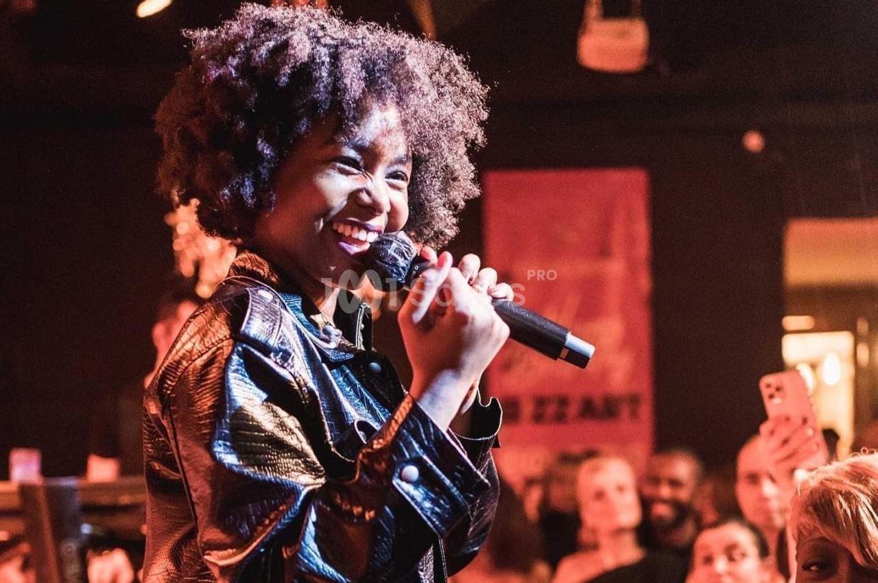 Une chanteuse souriante tenant un micro, sur scène devant un public attentif dans une ambiance chaleureuse.