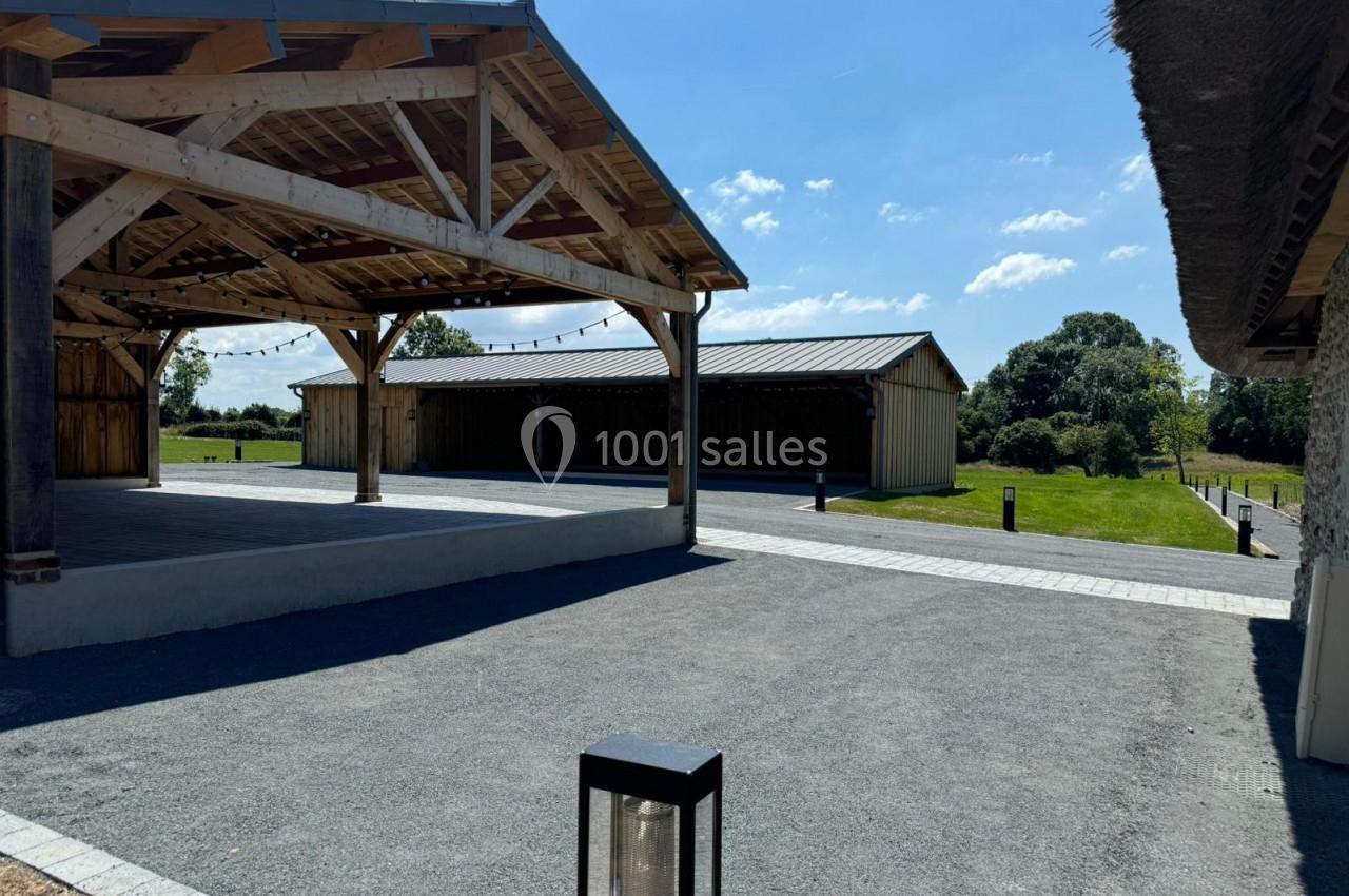 Structure en bois ouverte avec toit, espace pavé et bâtiment annexe en arrière-plan sous un ciel dégagé.