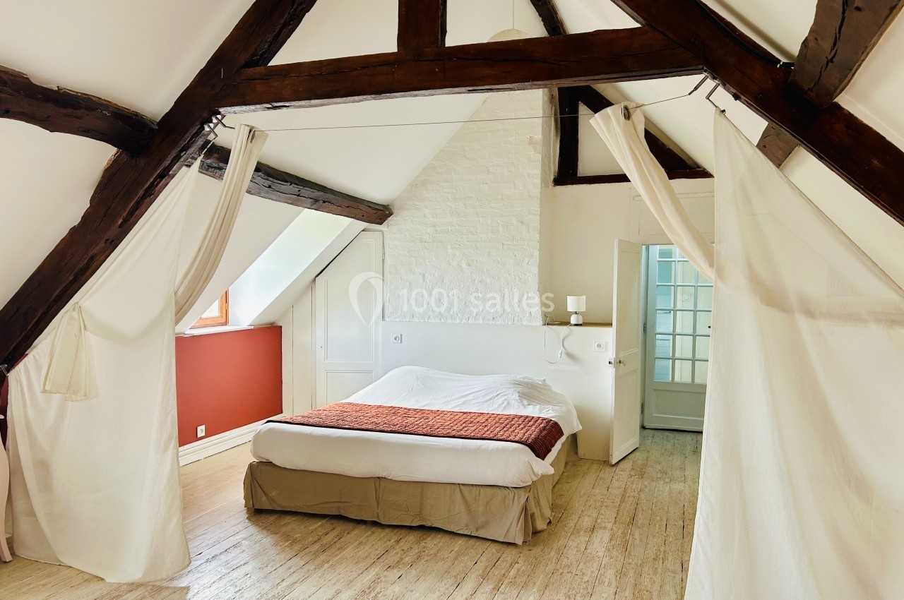 Chambre mansardée avec poutres apparentes, lit double, sol en bois clair et murs blancs, éclairée par une fenêtre.