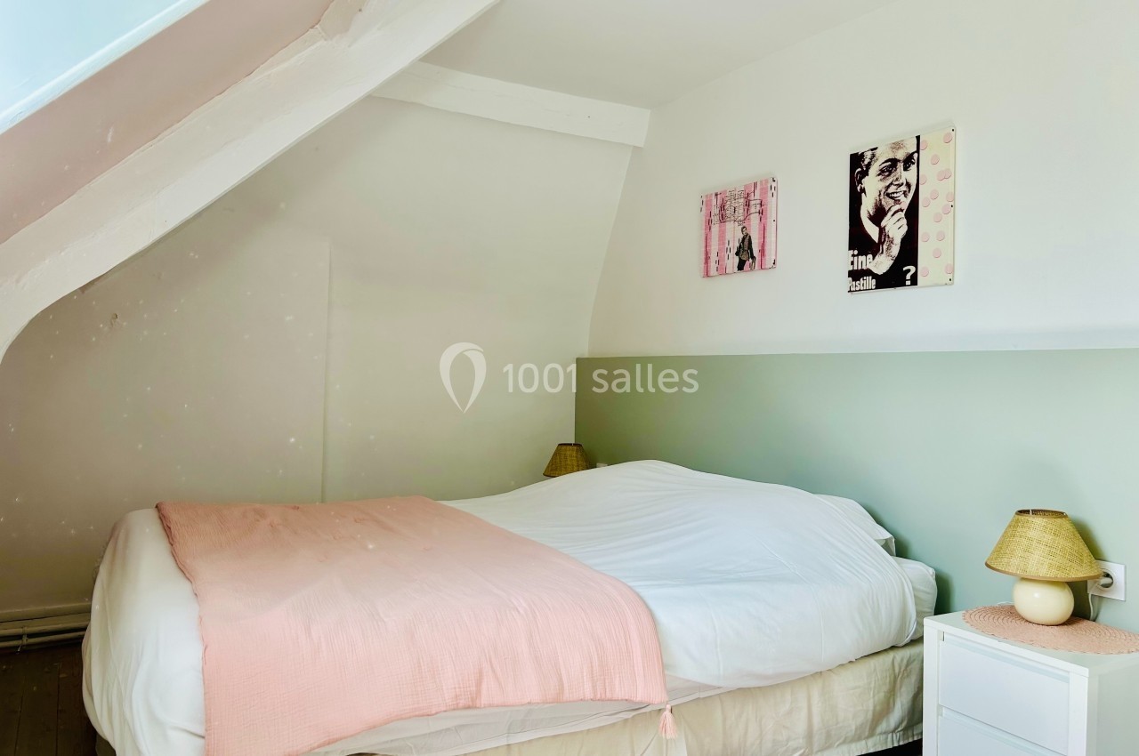 Chambre mansardée lumineuse avec lit double, couvre-lit rose, tables de chevet et décorations murales.