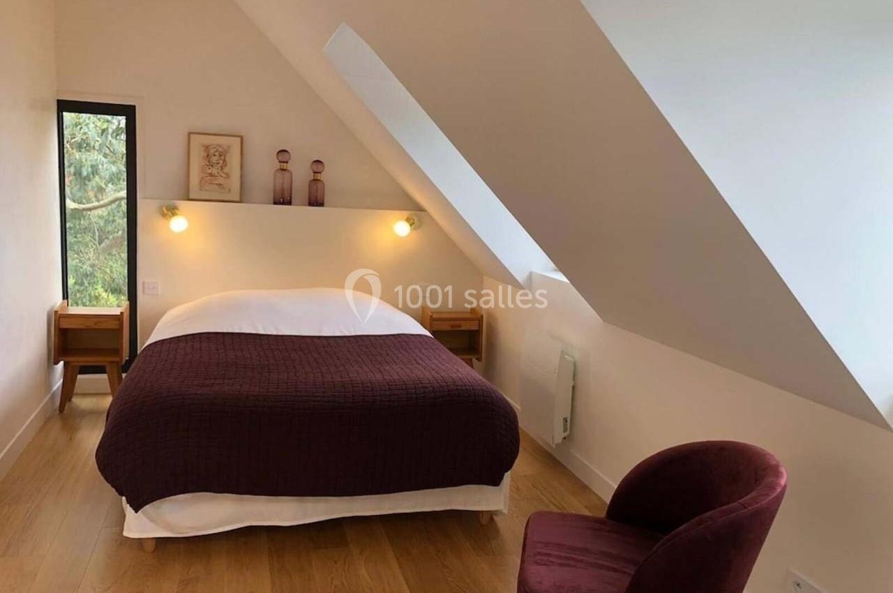 Chambre mansardée avec lit double, couvre-lit violet, fauteuil assorti, deux tables de chevet et fenêtre donnant sur des…