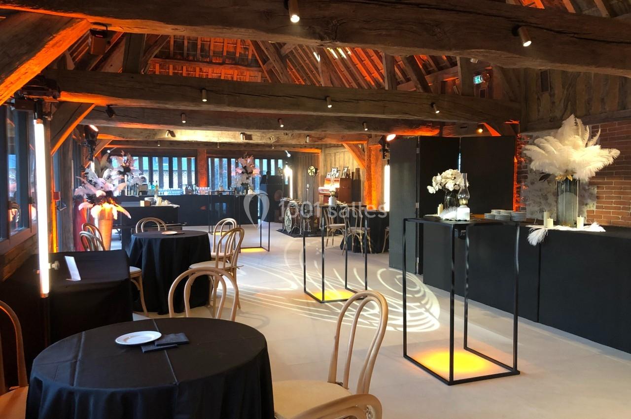 Salle de réception avec poutres apparentes, tables rondes, chaises en bois et décorations lumineuses et florales.