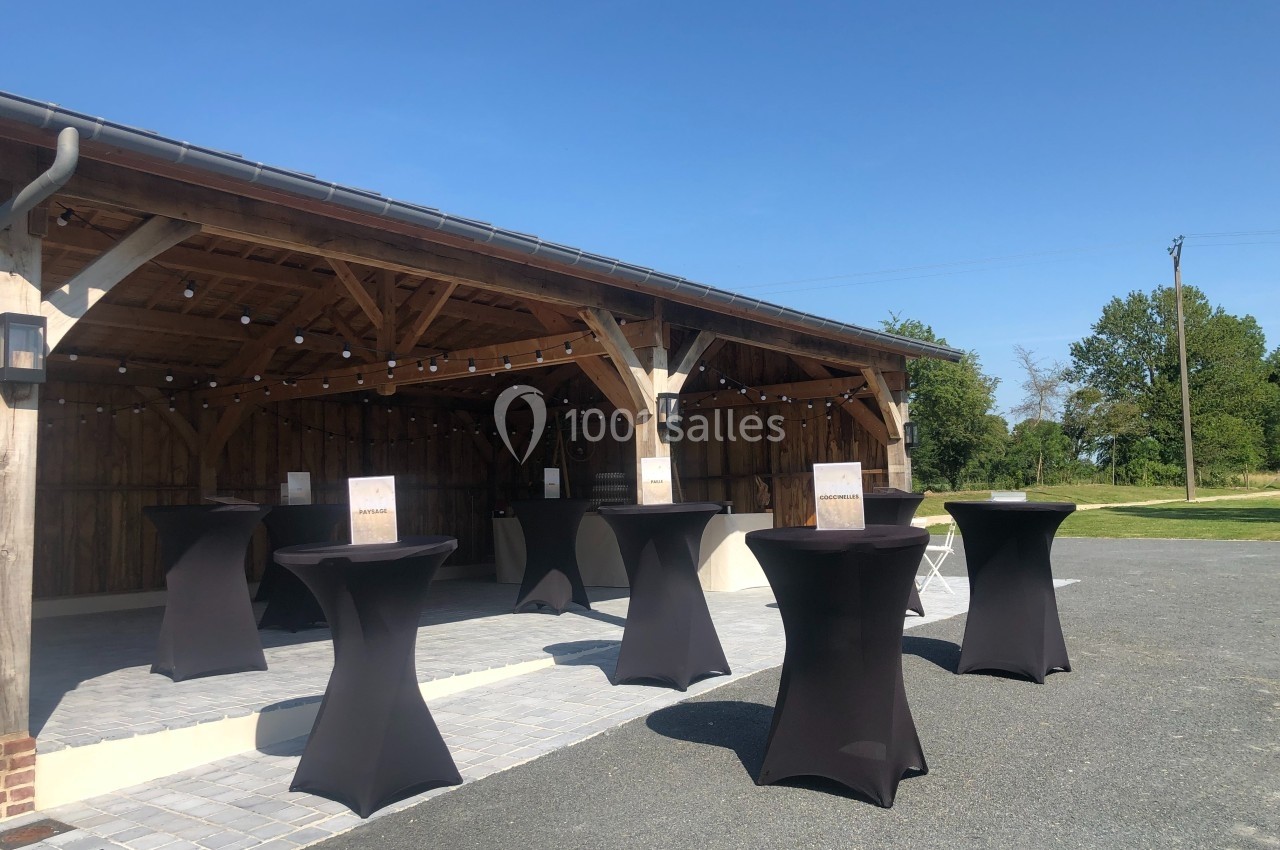 Espace extérieur avec tables hautes noires sous une structure en bois, éclairée par des guirlandes lumineuses.