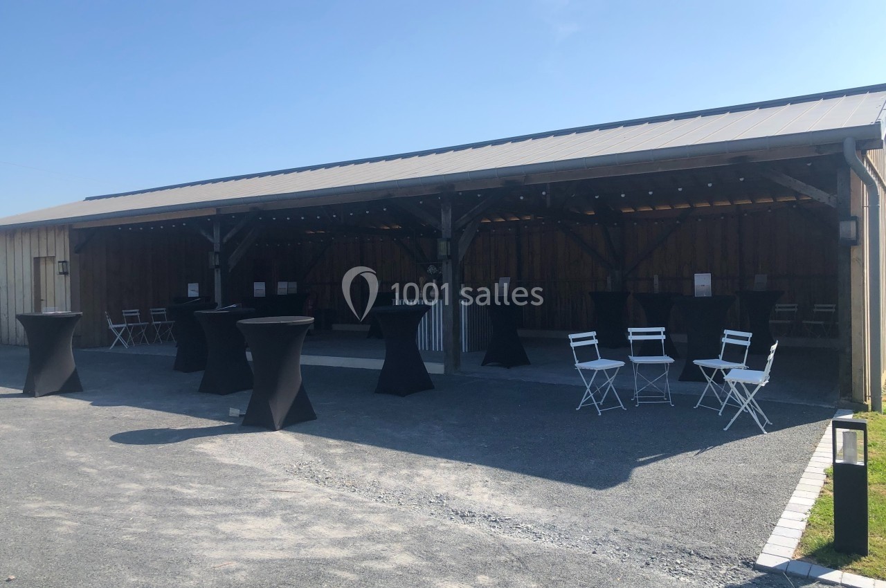 Abri en bois ouvert avec tables hautes noires et chaises blanches disposées sur un sol en gravier, sous un ciel dégagé.