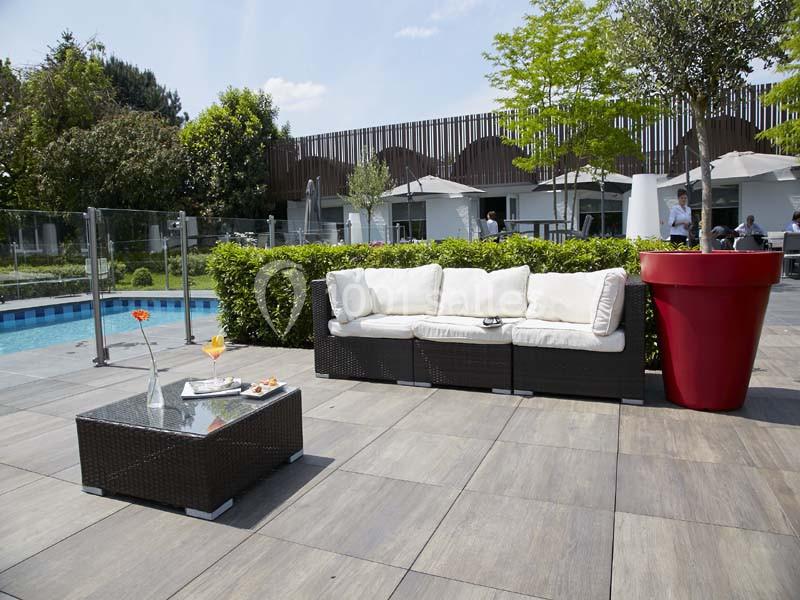 Location salle Noyelles-Godault (Pas-de-Calais) - Novotel Lens Noyelles #12 Salon de jardin avec canapé et table basse près d'une piscine, entouré de verdure et de parasols.