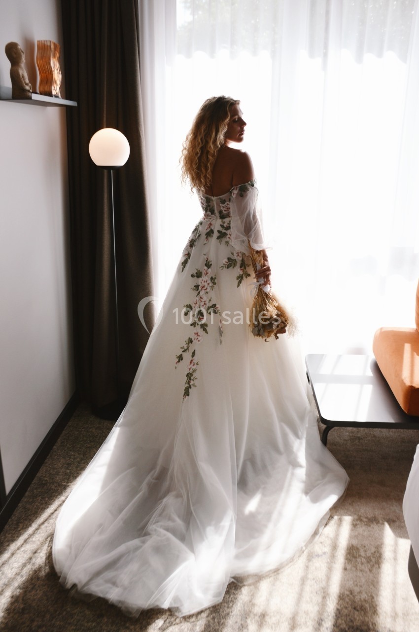 Location salle Noyelles-Godault (Pas-de-Calais) - Novotel Lens Noyelles #16 Femme en robe de mariée blanche à motifs floraux, tenant un bouquet, debout près d'une fenêtre lumineuse.
