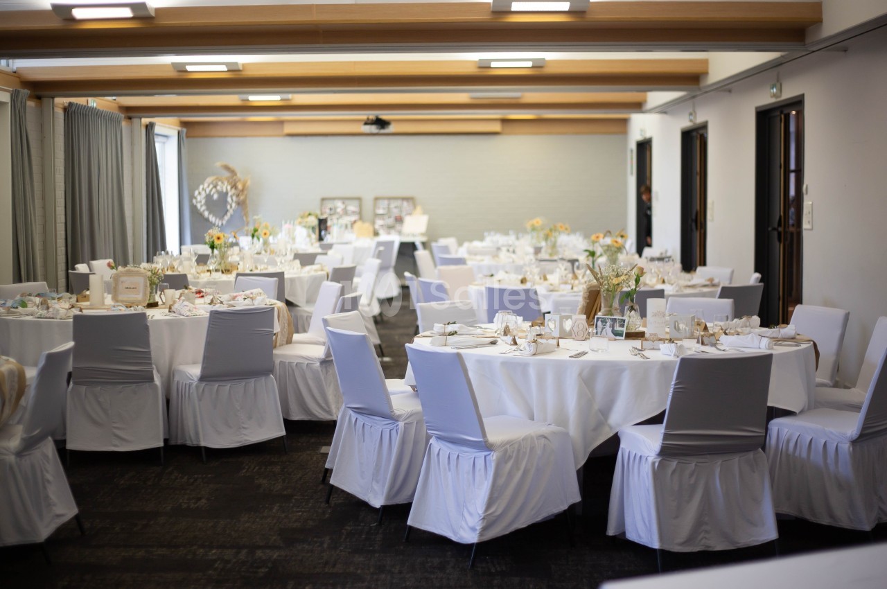 Salle de réception décorée avec des tables rondes, nappes blanches, chaises houssées et arrangements floraux.
