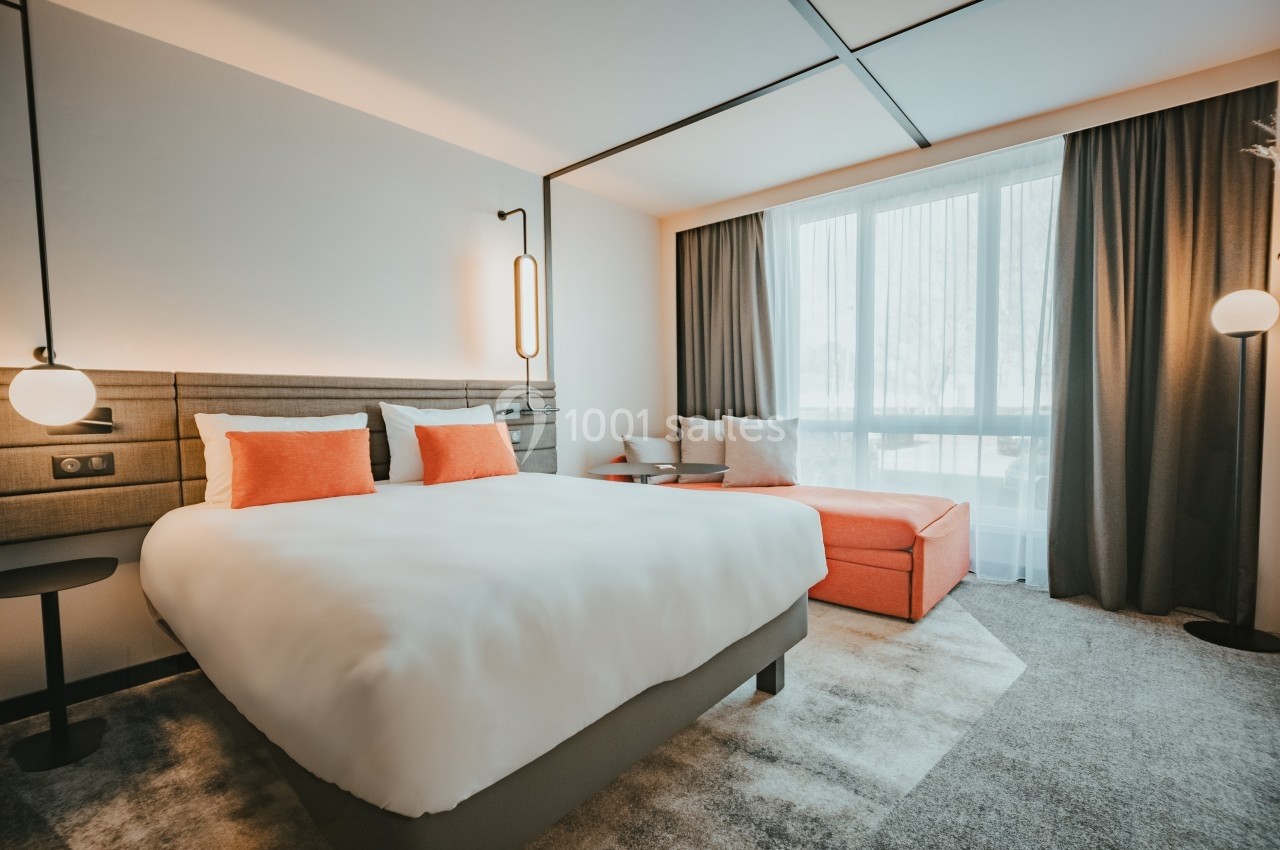 Location salle Noyelles-Godault (Pas-de-Calais) - Novotel Lens Noyelles #17 Chambre d'hôtel lumineuse avec un lit double, coussins orange, canapé assorti et grande fenêtre avec rideaux.