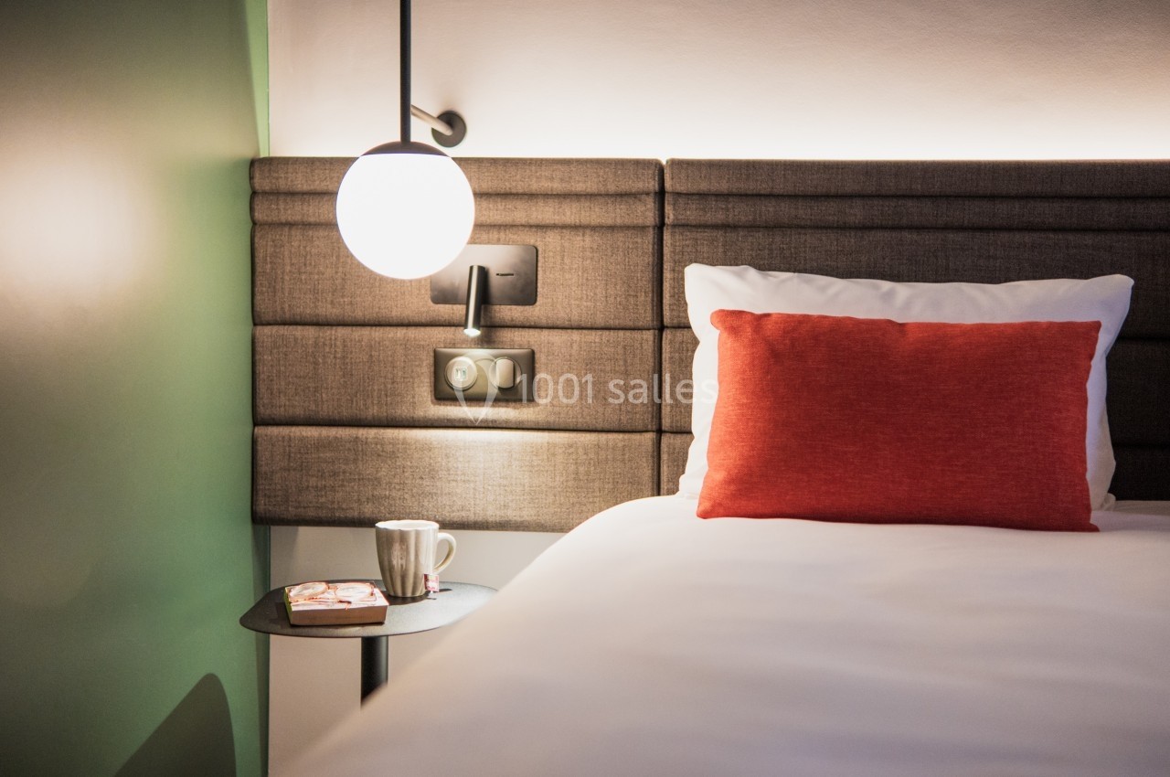 Location salle Noyelles-Godault (Pas-de-Calais) - Novotel Lens Noyelles #18 Lit avec tête de lit rembourrée, coussin rouge, lampe suspendue et table de chevet avec tasse et livre.