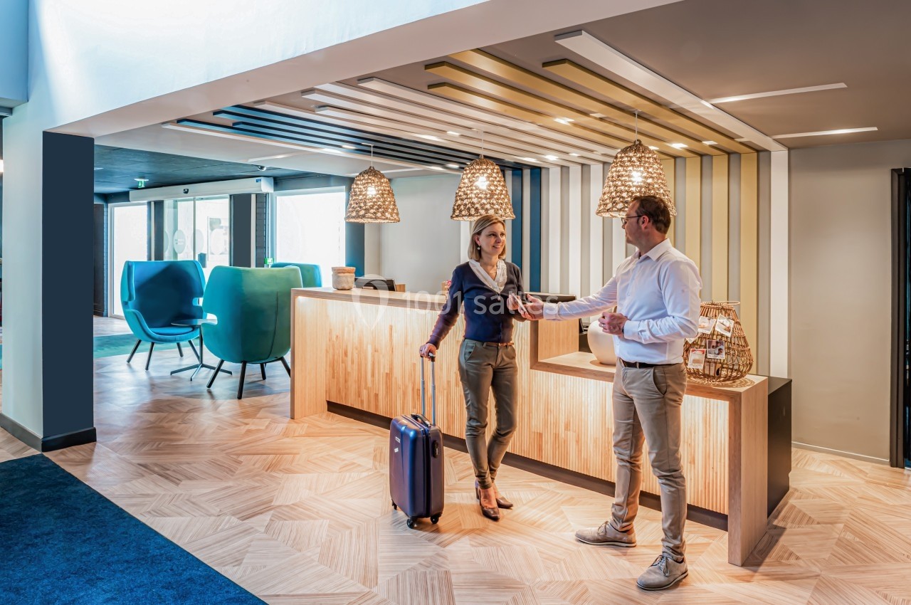 Location salle Noyelles-Godault (Pas-de-Calais) - Novotel Lens Noyelles #21 Un homme accueille une femme avec une valise dans un hall d'hôtel moderne et lumineux.