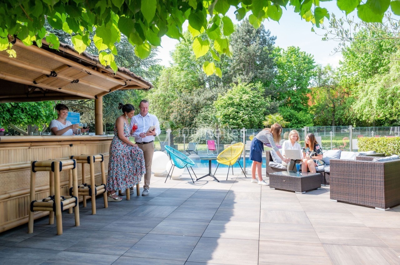 Location salle Noyelles-Godault (Pas-de-Calais) - Novotel Lens Noyelles #3 Personnes discutant et se détendant sur une terrasse près d'un bar en bois, avec piscine et végétation en arrière-plan.