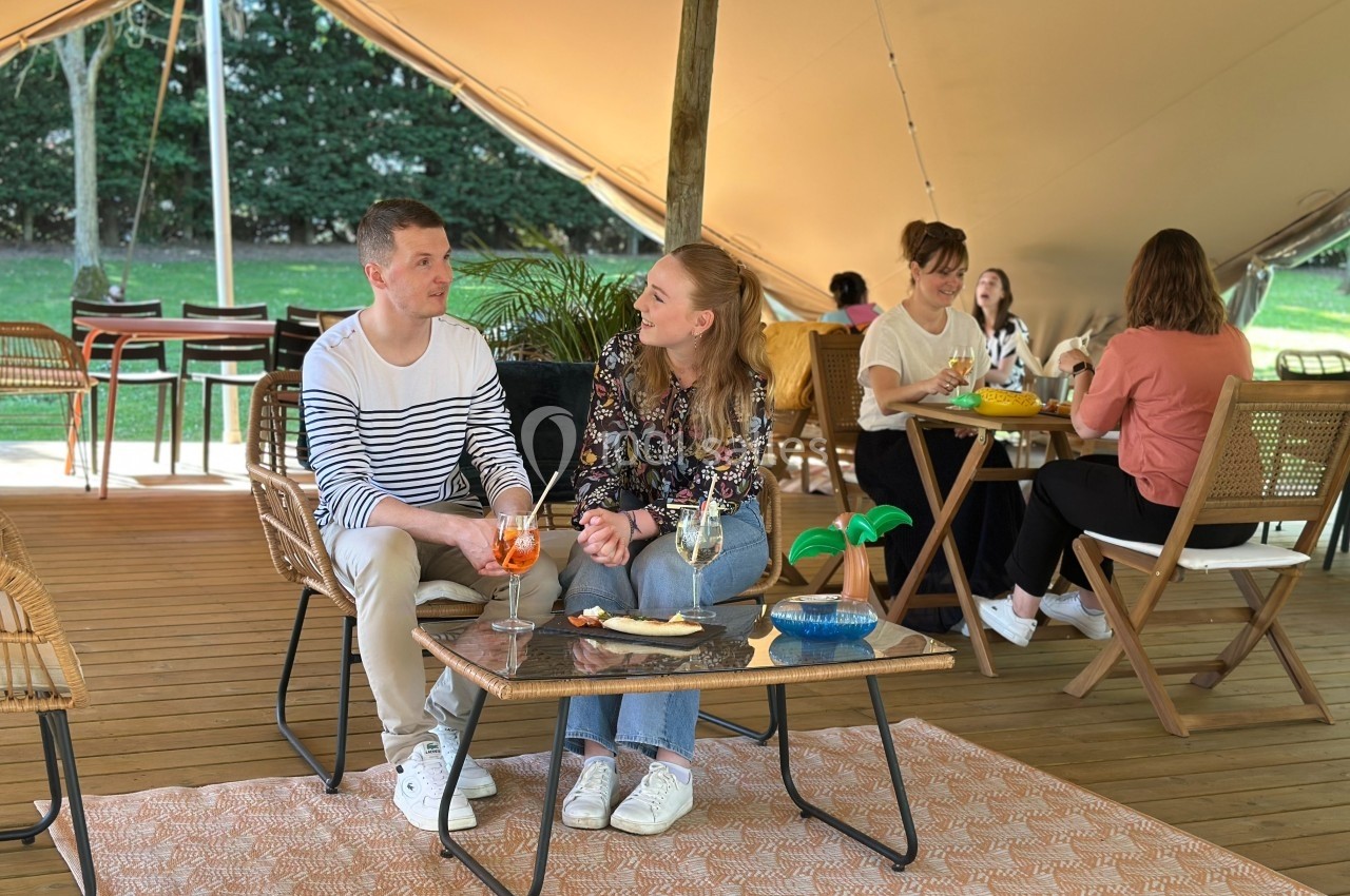 Location salle Noyelles-Godault (Pas-de-Calais) - Novotel Lens Noyelles #4 Deux personnes discutent autour d'une table avec des boissons sous une tente, d'autres personnes assises en arrière-plan.