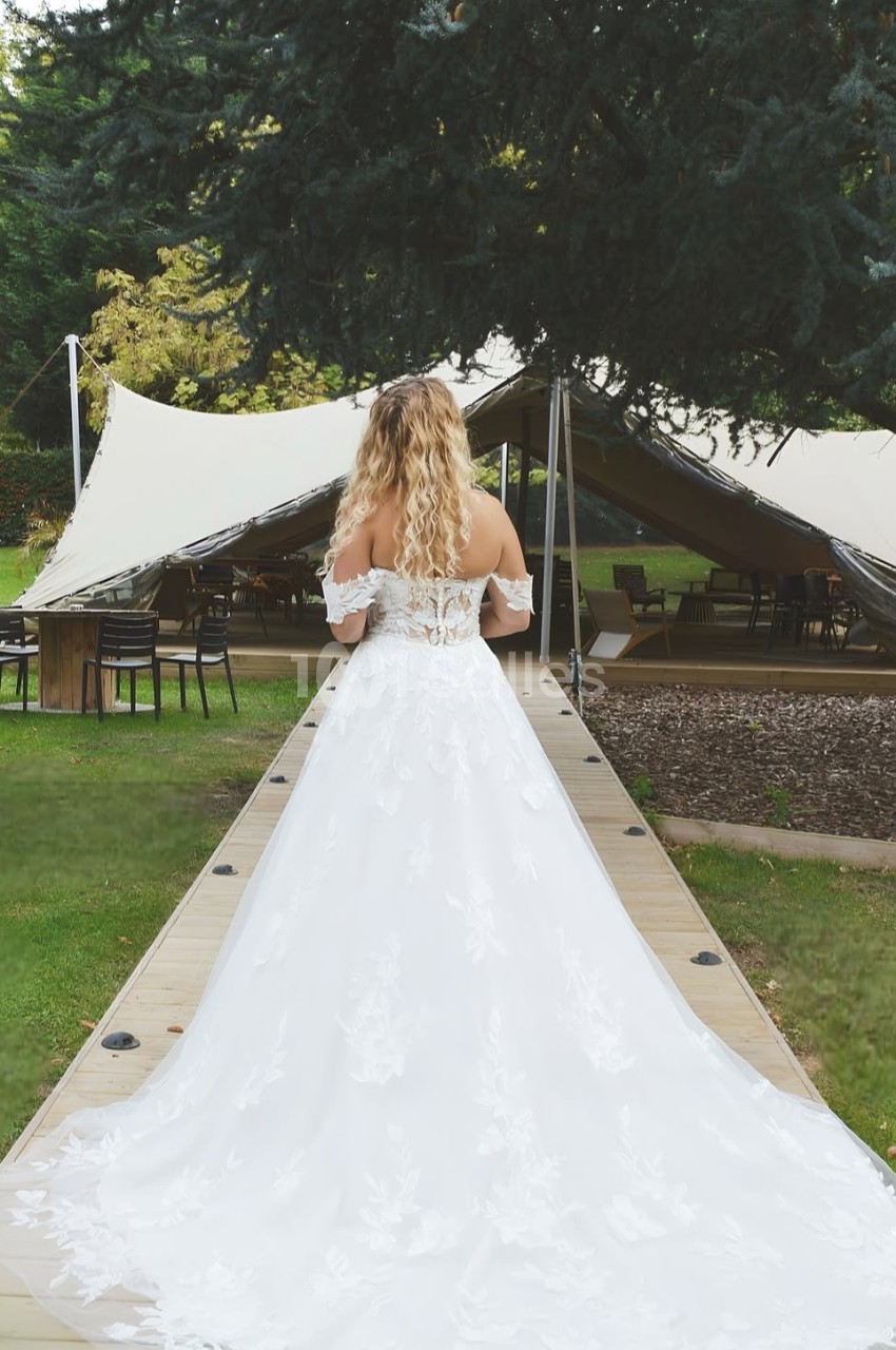 Femme en robe de mariée blanche vue de dos, marchant sur une passerelle en bois vers une tente dans un jardin.