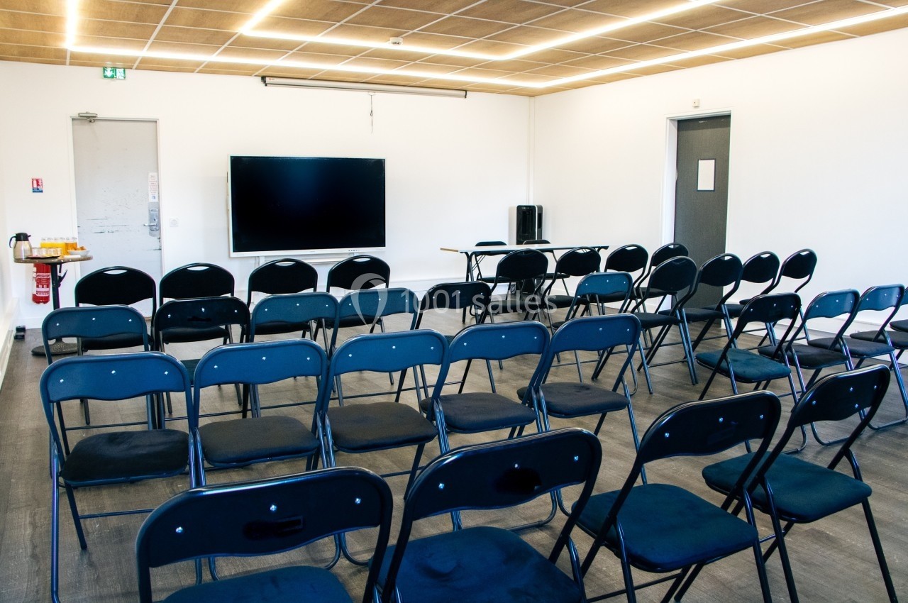 Salle de réunion avec des rangées de chaises noires, un écran mural et un plafond en bois clair.
