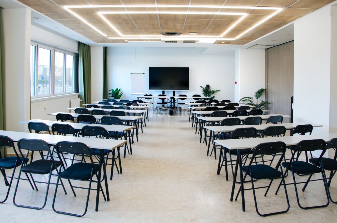 Salle de conférence lumineuse avec des rangées de tables et chaises noires, un écran et un tableau blanc à l'avant.