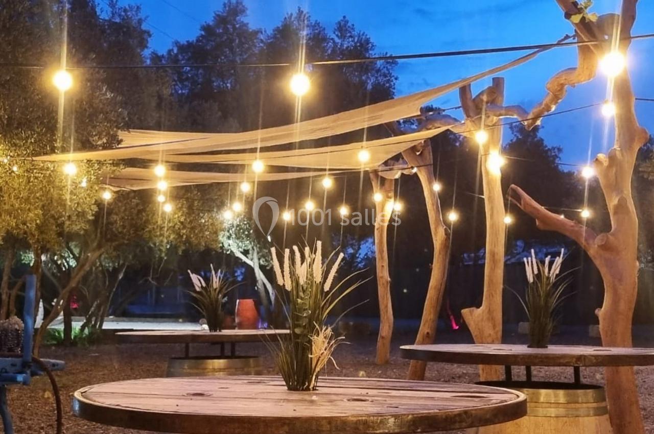 Guirlandes lumineuses suspendues au-dessus de tables en bois dans un espace extérieur décoré de plantes et d'arbres.