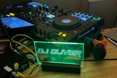 Table de mixage avec platines, microphones et panneau lumineux indiquant ’DJ Olivier’ sur une table en bois.