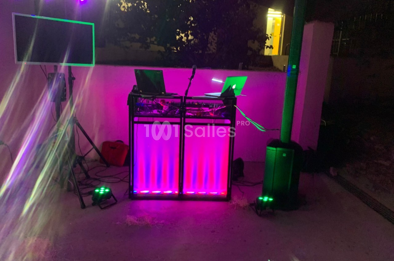 Poste de DJ éclairé par des lumières colorées, avec des équipements audio et un écran dans un espace extérieur sombre.