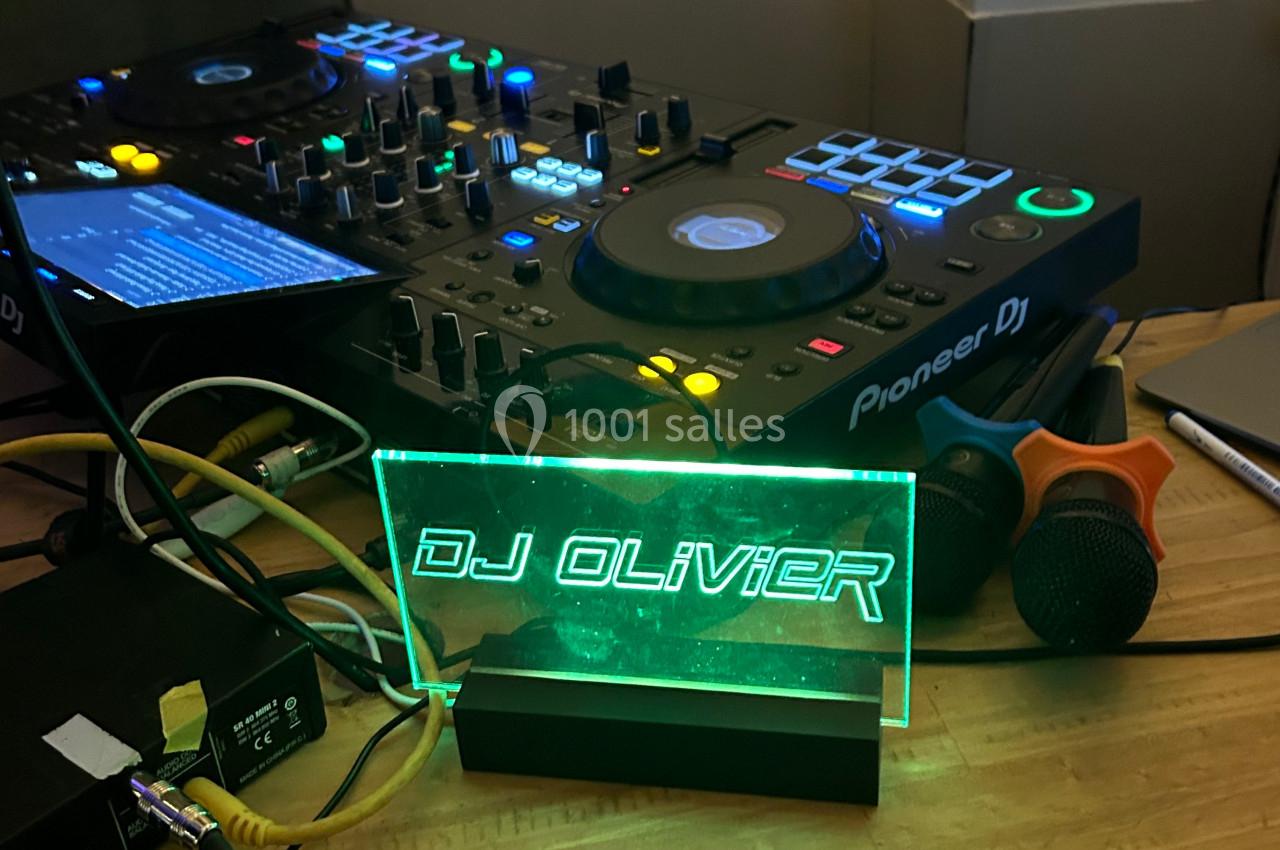 Table de mixage avec platines, microphones et panneau lumineux indiquant ’DJ Olivier’ sur une table en bois.