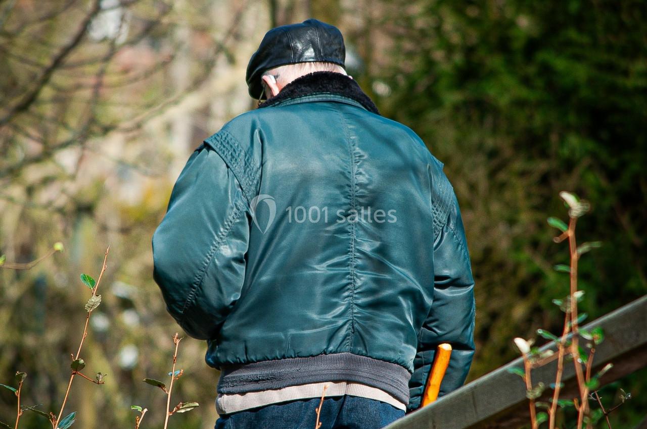 Un homme âgé portant une veste verte et une casquette noire marche avec une canne dans un environnement extérieur.