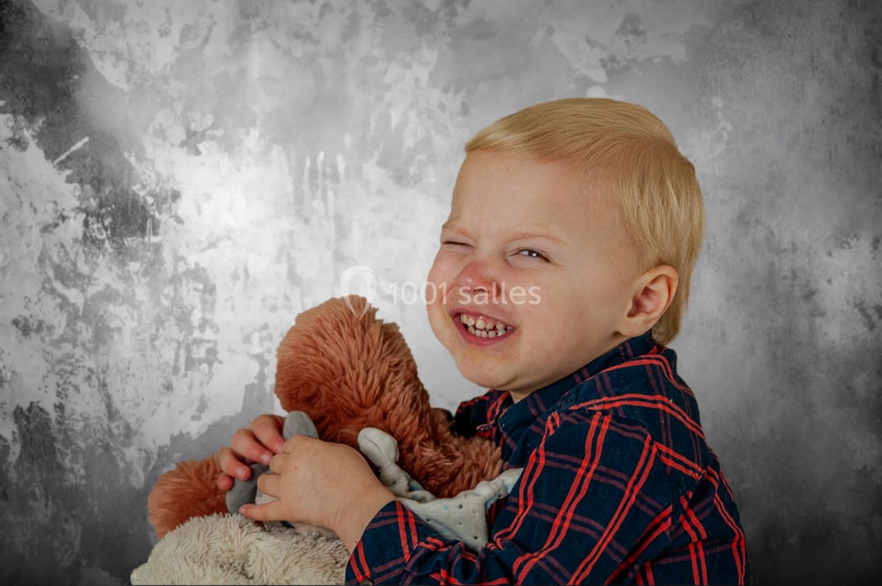 Un enfant souriant, vêtu d'une chemise à carreaux, serre une peluche contre un fond gris texturé.