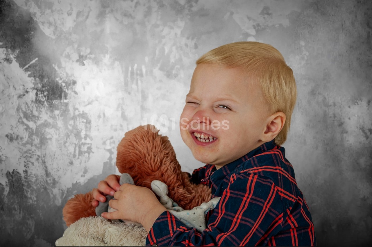 Un enfant souriant, vêtu d'une chemise à carreaux, serre une peluche contre un fond gris texturé.