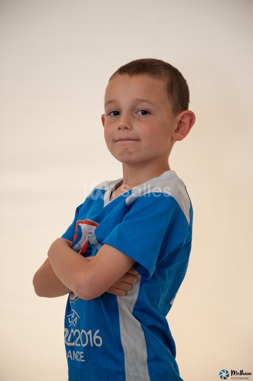 Un enfant en t-shirt bleu croise les bras devant un fond clair, regardant l'objectif avec une expression confiante.