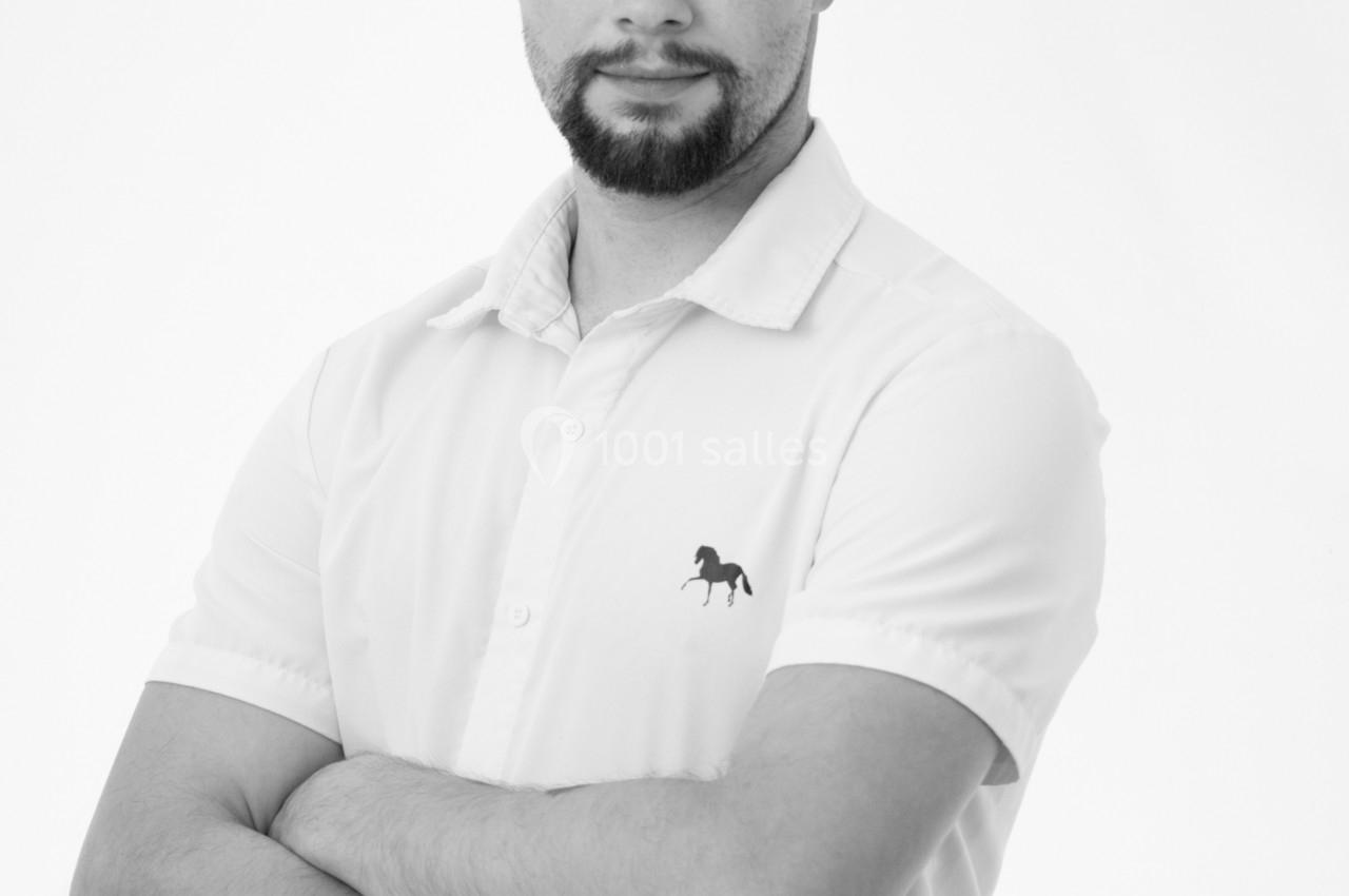 Homme debout, bras croisés, portant une chemise blanche avec un motif de cheval, fond blanc.