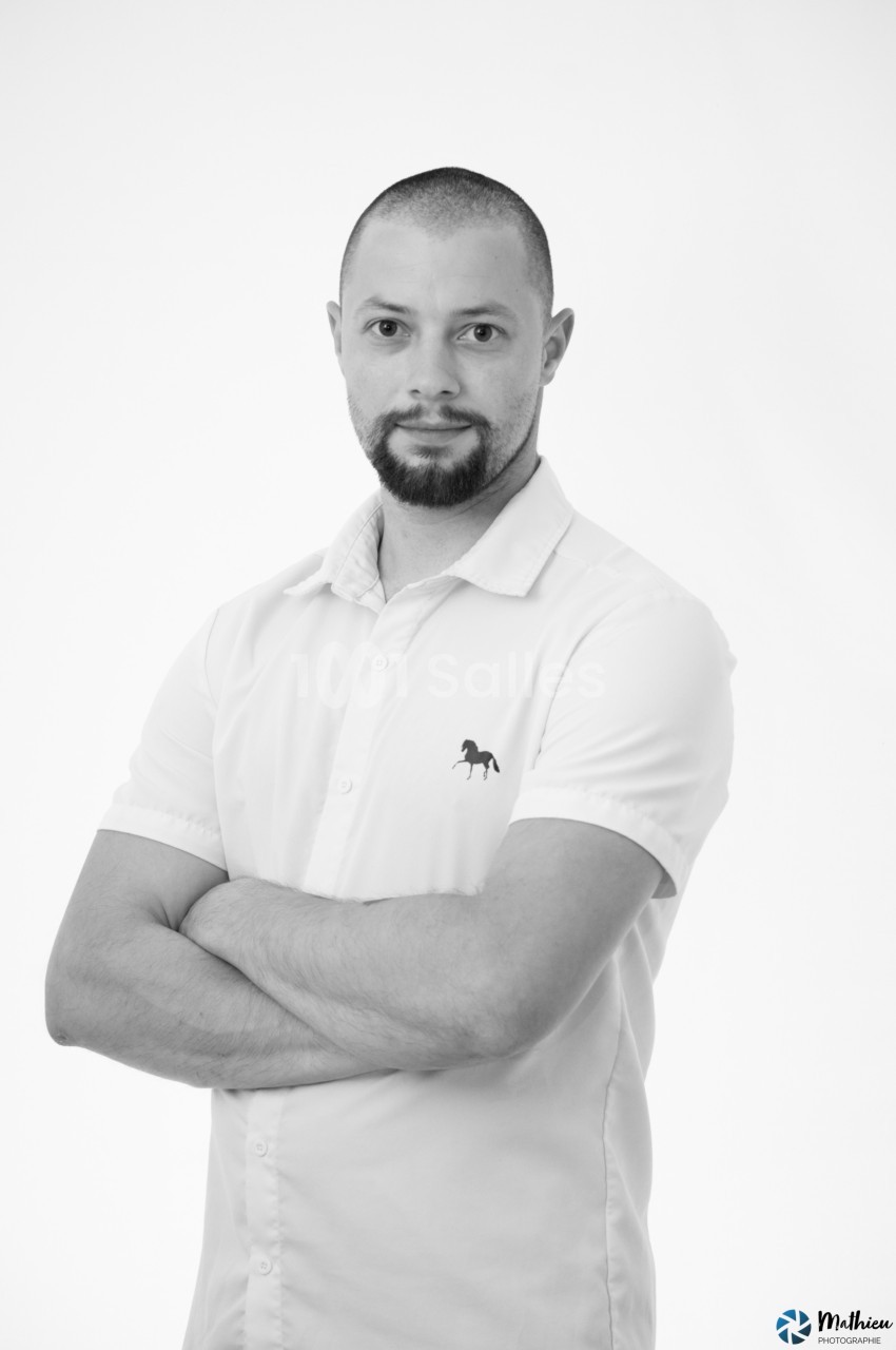 Homme debout, bras croisés, portant une chemise blanche avec un motif de cheval, fond blanc.