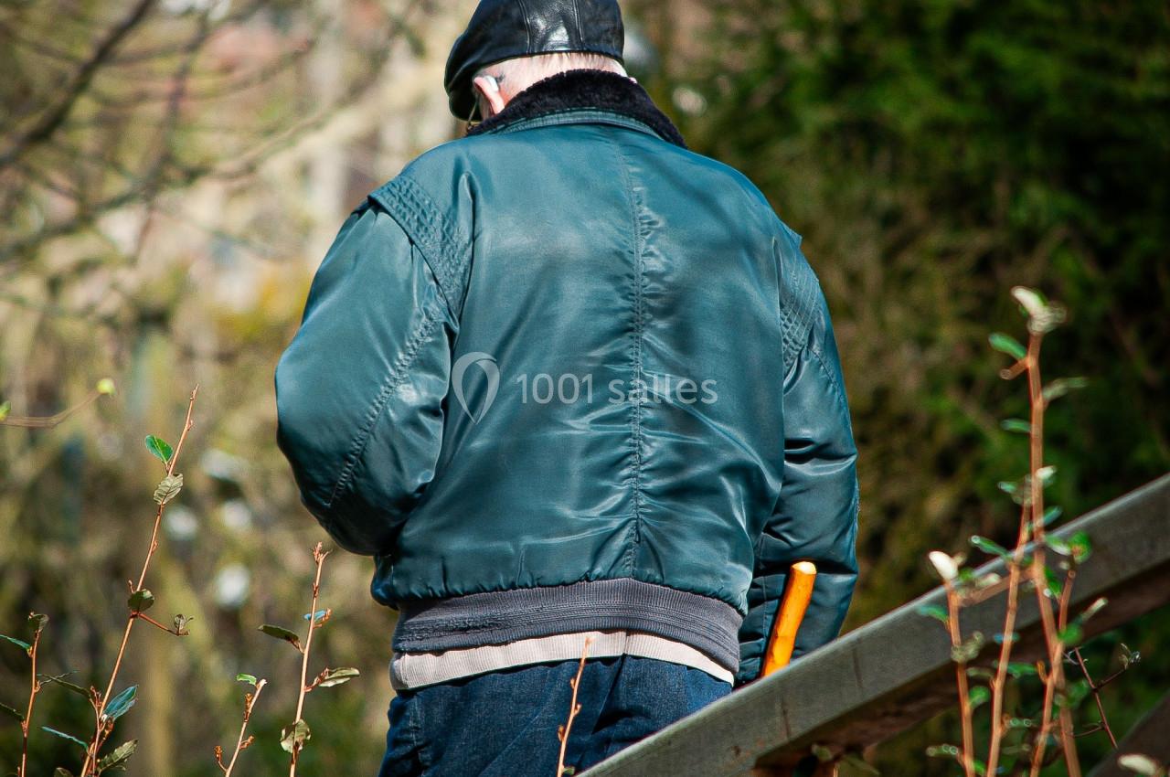 Un homme portant une veste verte et un béret marche sur un sentier boisé, appuyé sur une canne.
