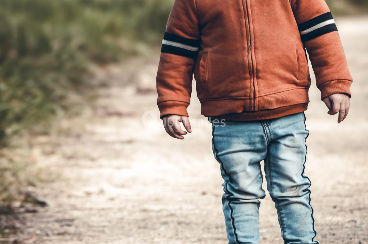 Un enfant souriant en sweat orange regarde un petit drone posé sur un chemin en plein air.