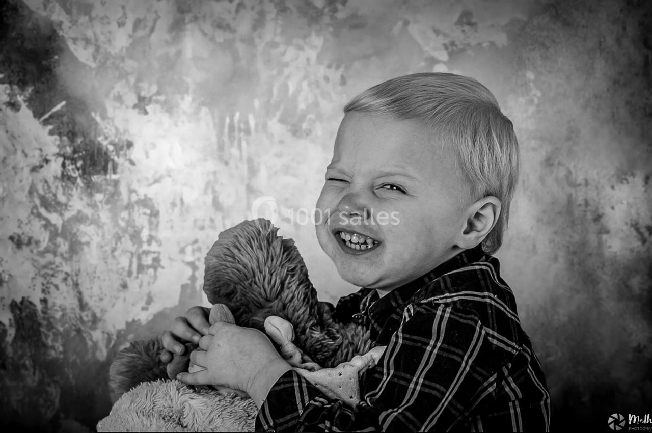 Un enfant souriant en noir et blanc, tenant un ours en peluche devant un fond texturé.