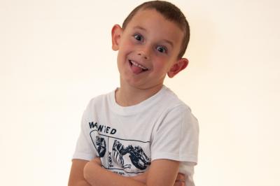 Un enfant souriant en t-shirt blanc pose avec une main sous le menton sur un fond clair.