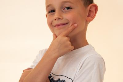 Un enfant souriant en t-shirt blanc pose avec une main sous le menton sur un fond clair.