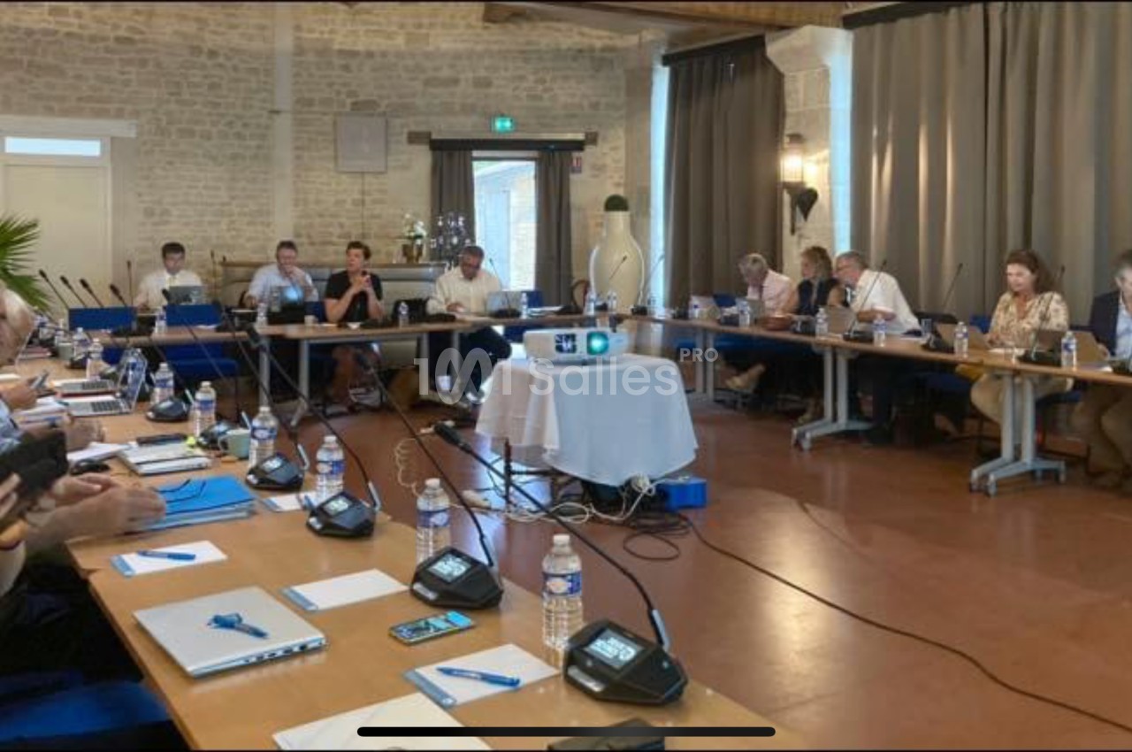 Réunion autour d'une grande table en U, avec des participants, des micros et un projecteur au centre.