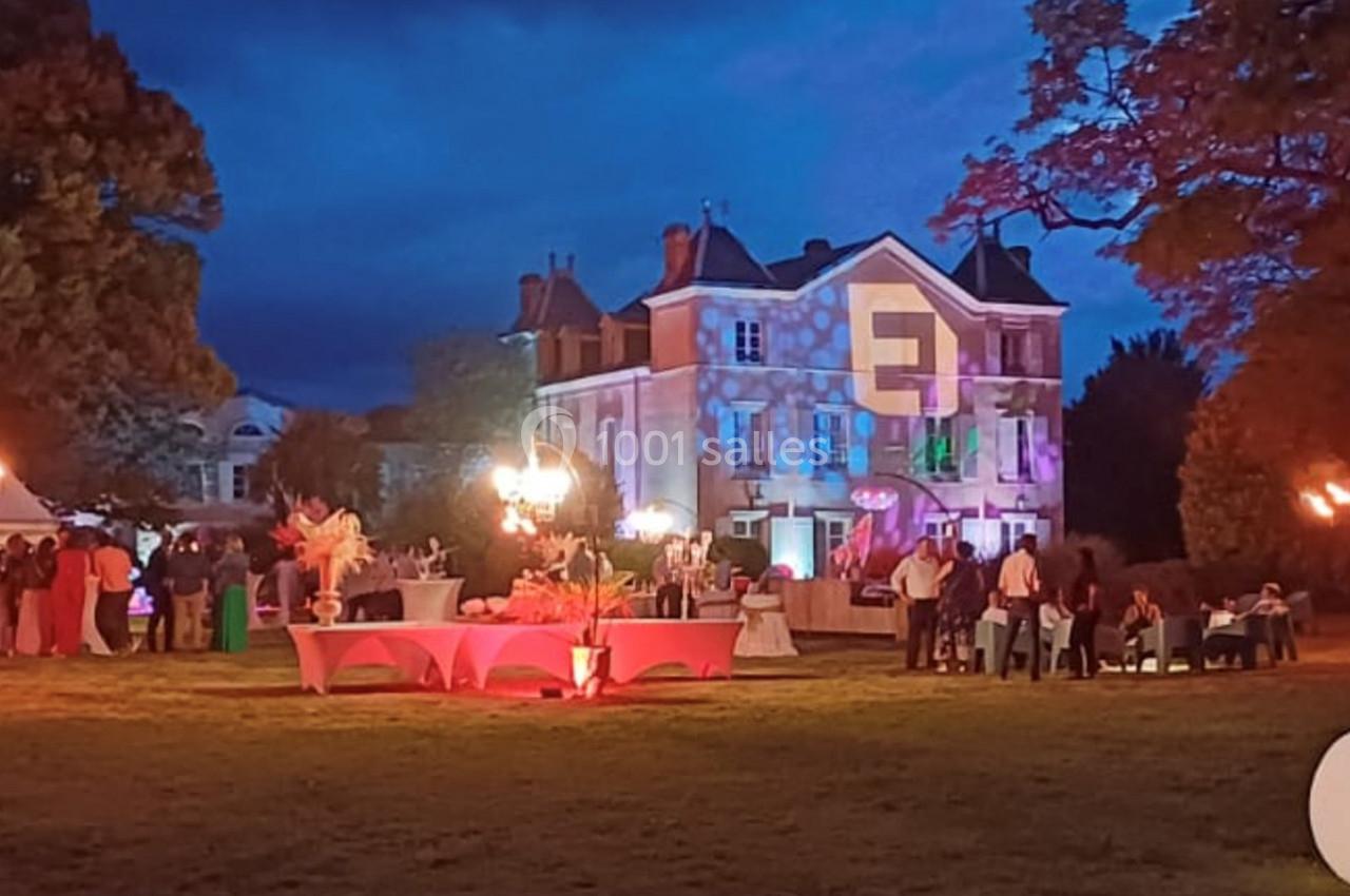 Manoir éclairé de nuit avec des lumières colorées, tables dressées et invités rassemblés dans un jardin.