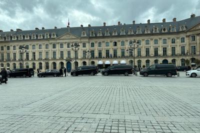 Façade d'un bâtiment historique avec des voitures noires alignées sur une place pavée sous un ciel nuageux.